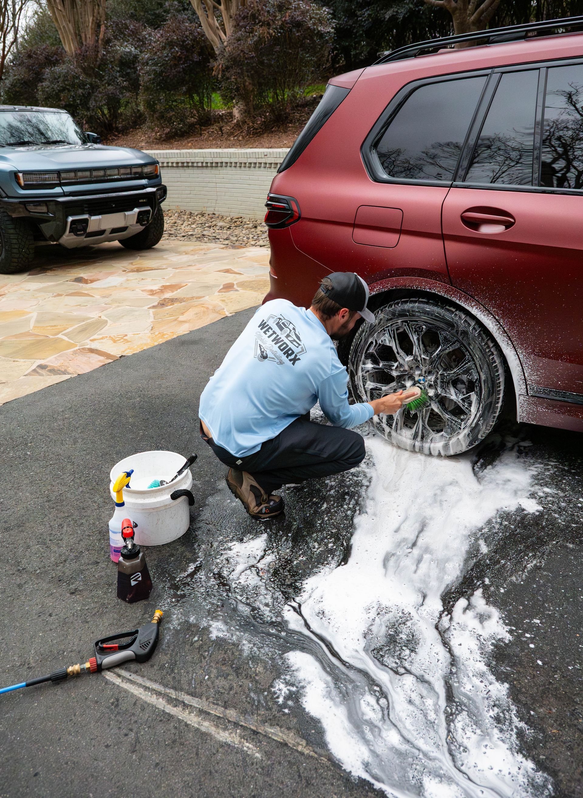 Man washes a red SUV's wheel on a driveway. Soapy water surrounds him. Another car is visible behind.
