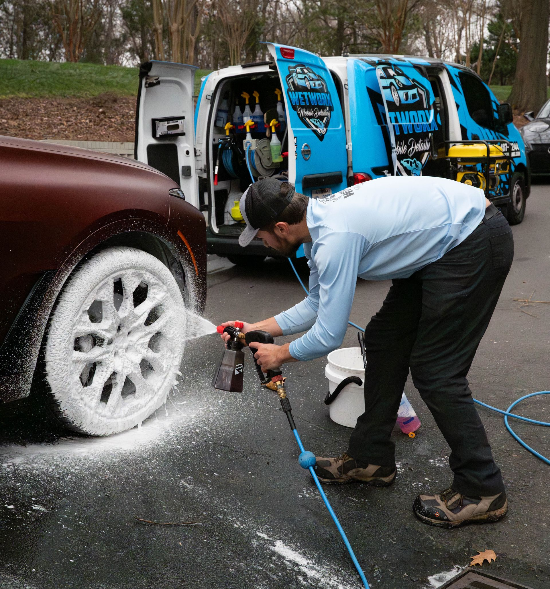 Man washes car wheel with foam, parked next to a van with cleaning supplies.