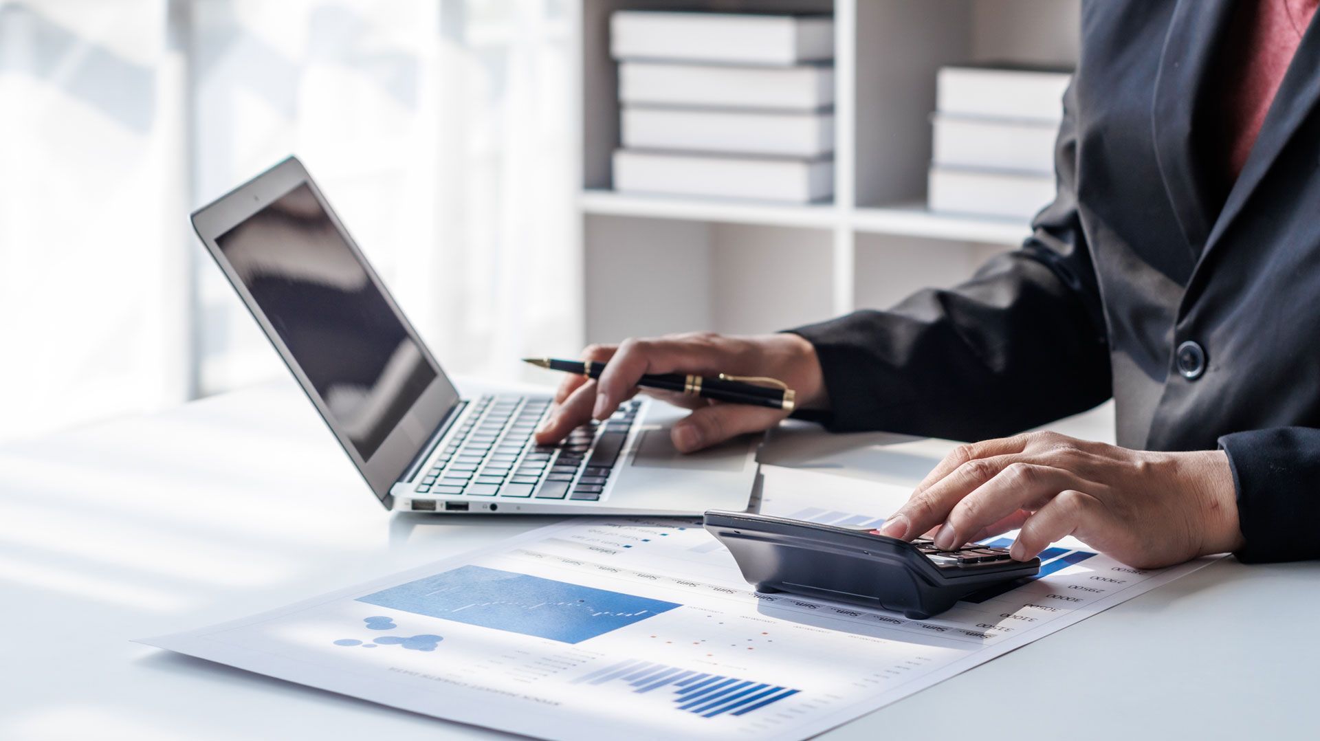 Person in suit using a laptop and calculator, analyzing financial charts at a desk.