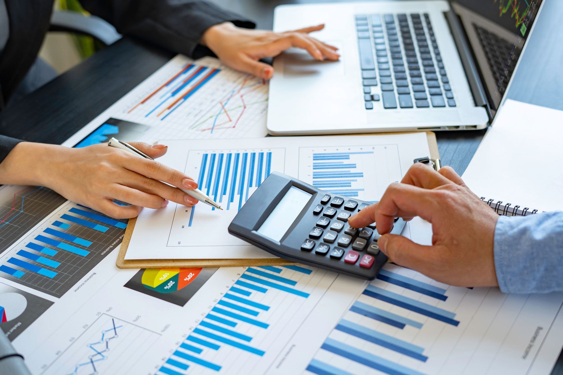 Hands analyzing financial reports with a calculator and laptop on a desk.