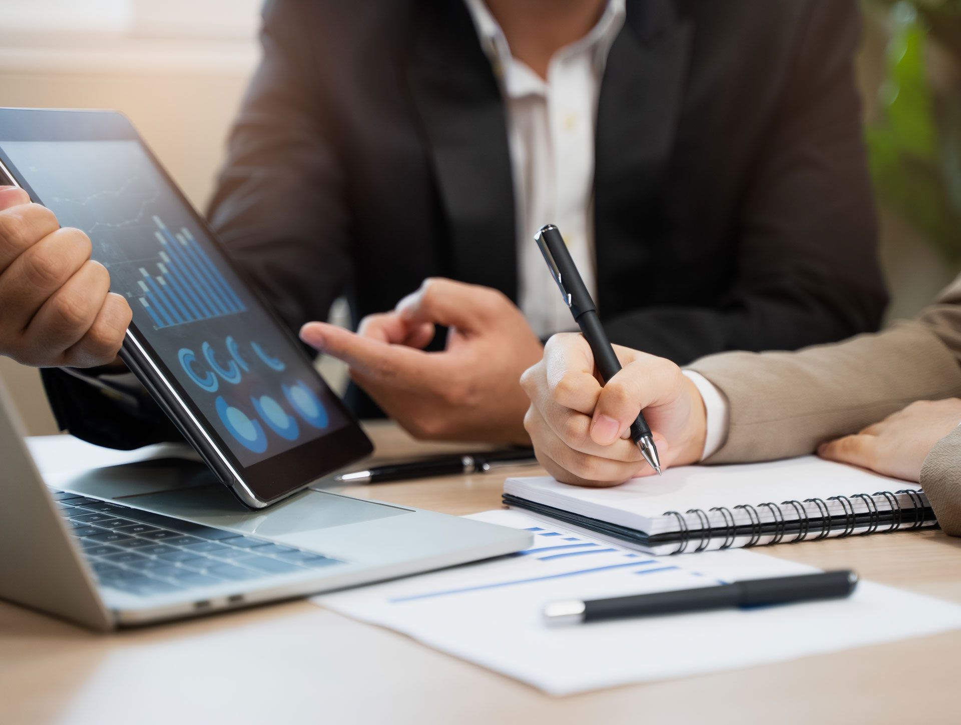 People in business attire reviewing data on a tablet, taking notes with a pen, and using a laptop at a table.