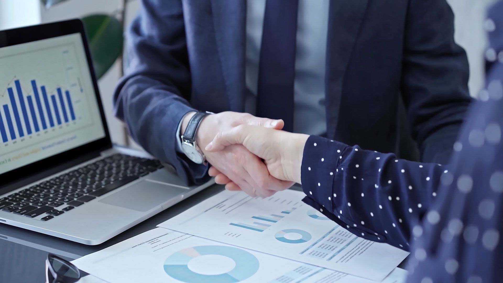 Two businesspeople shaking hands over documents and a laptop with graphs.