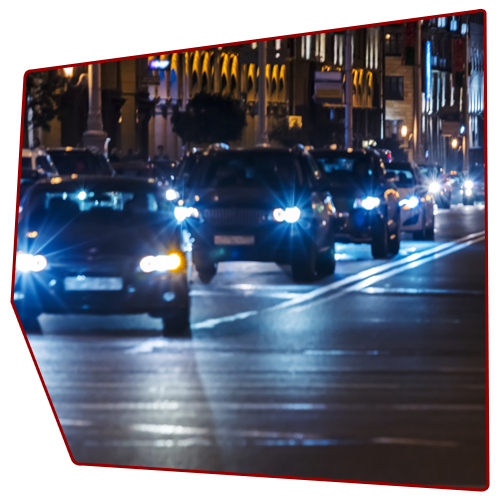 Cars driving on a city street at night, with bright headlights and city lights in the background.