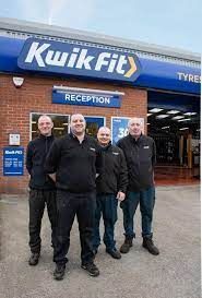 A group of men are standing in front of a kwik fit tire shop.