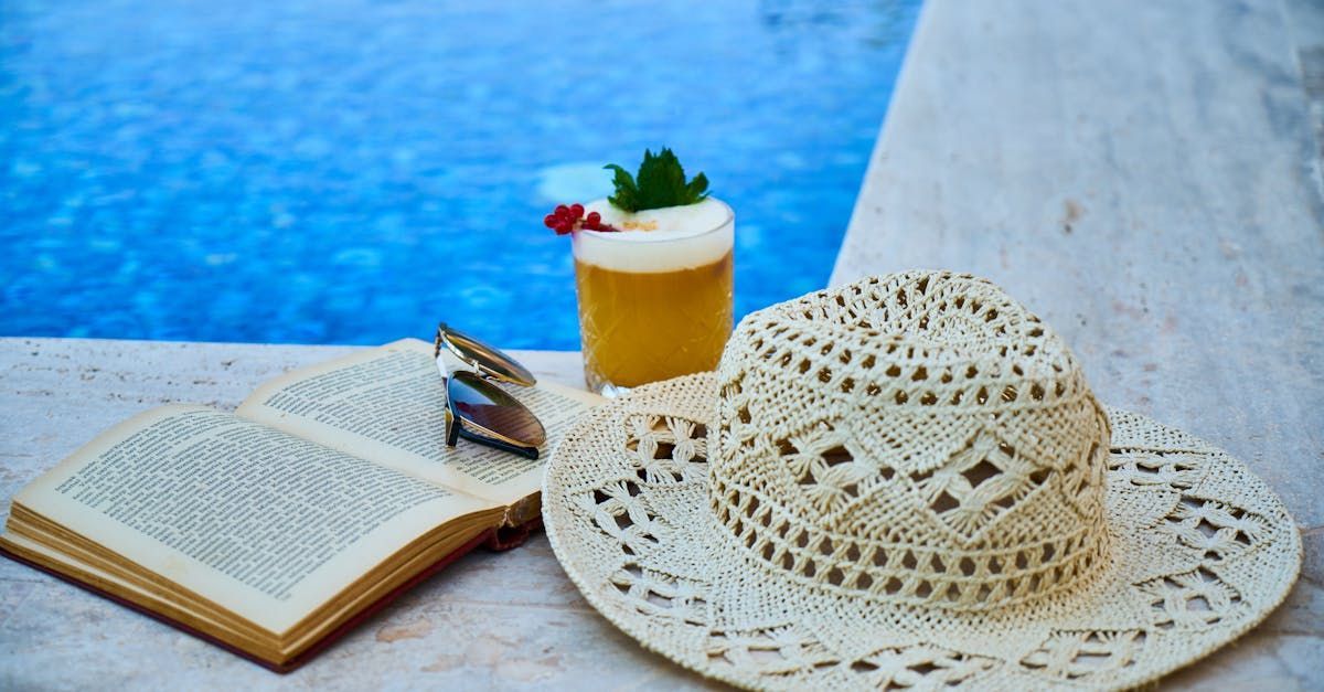 A straw hat , a book , and a drink are sitting next to a swimming pool.