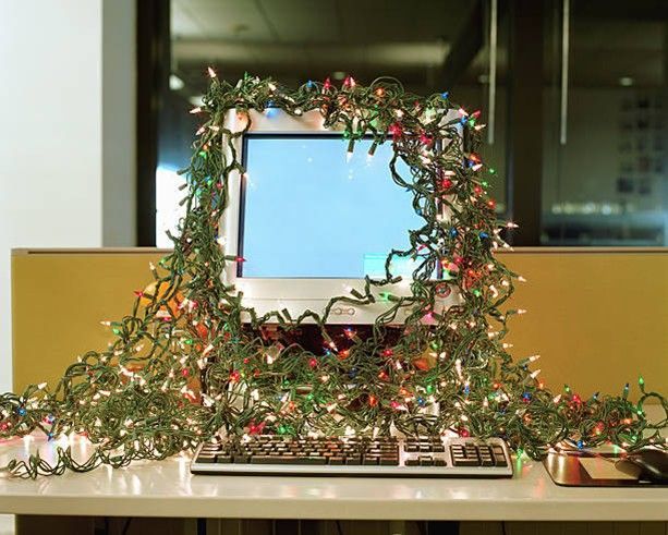 Computer with Christmas lights draped around the screen and keyboard, on a desk in an office.