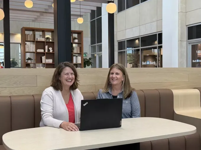 Two women smiling at a laptop, seated at a table in a modern office space.
