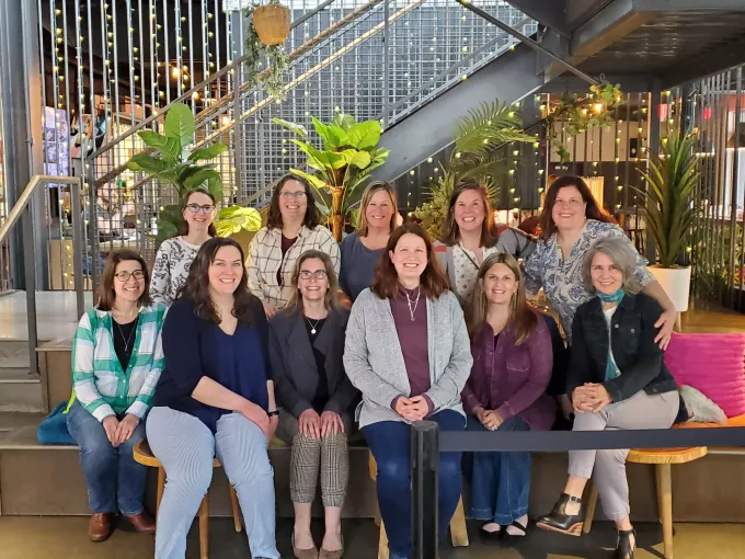 Group of 11 women smiling, posing together indoors near plants, stairs, and a metal structure.