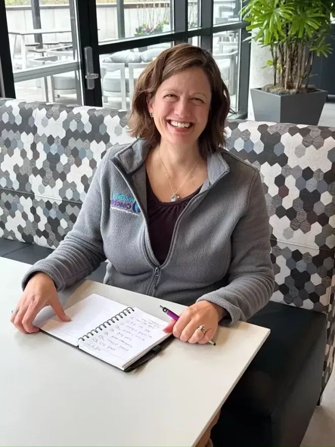 Woman smiles while sitting at a table with a notebook and pen. She wears a gray zip-up.