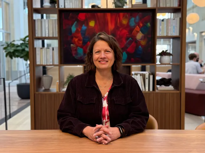Woman at a desk smiling, hands clasped.  Behind her is a bookshelf with books and art in a modern office space.