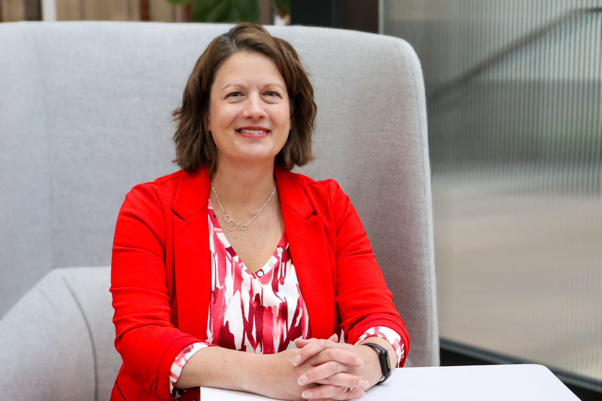 Woman in red blazer smiles, seated in a gray chair. Hands clasped, white table in front.