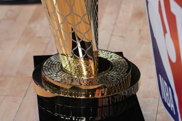 A close-up of a golden NBA championship trophy base resting on a polished wooden floor next to an NBA logo banner.