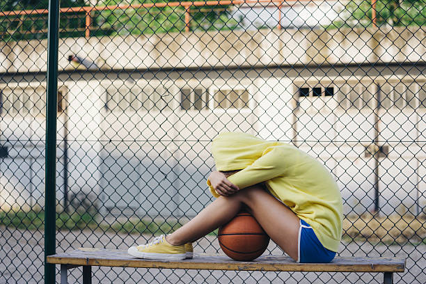 Person in yellow hoodie and shorts sits on bench, head down, knees up, next to a basketball, chain-link fence in background.