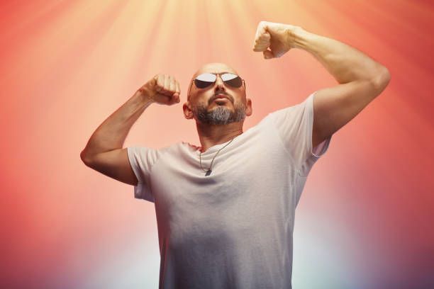 Bald man flexing, wearing sunglasses and white shirt, with arms raised. Red and yellow background.