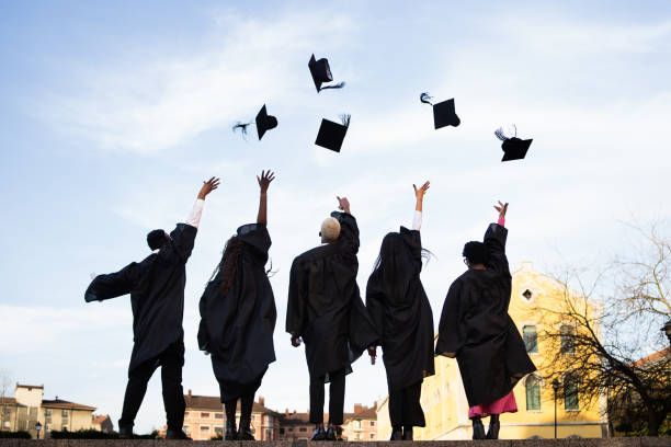 Five graduates in black gowns stand with their backs to the camera, tossing their mortarboards into a clear blue sky.