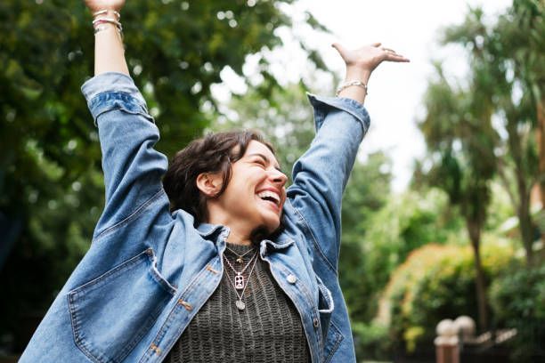 Woman with arms raised, smiling broadly outdoors, wearing a denim jacket.