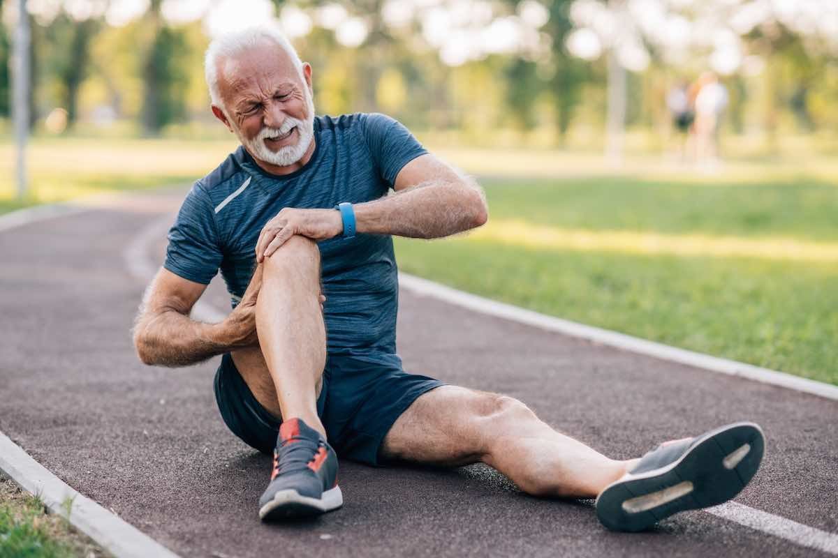 An older man is sitting on the ground holding his knee in pain.