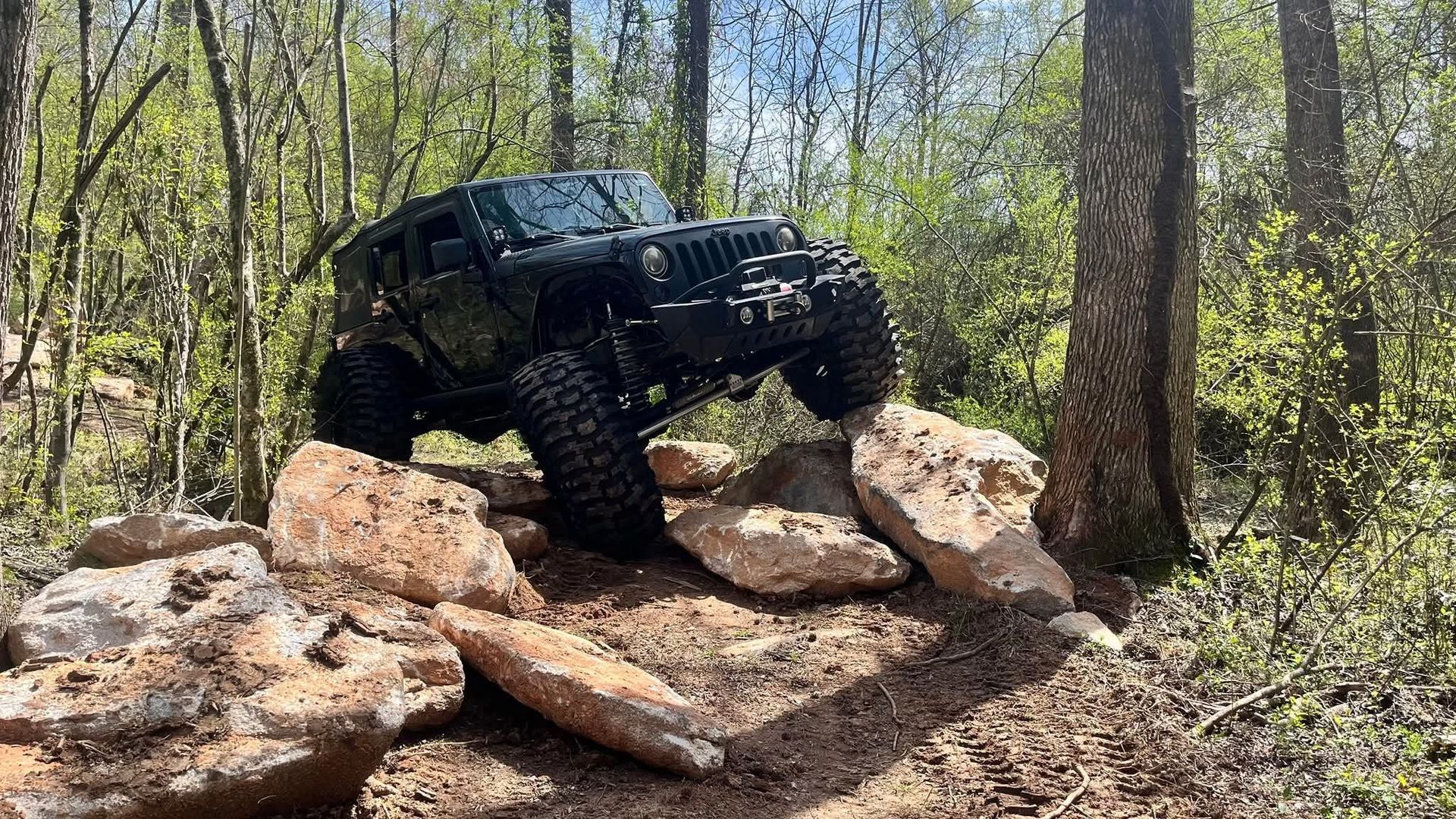 Black Jeep off-roading, climbing over rocks in a wooded area.