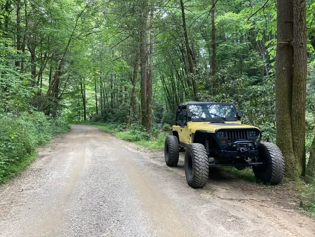 A yellow jeep is parked on the side of a dirt road in the woods.