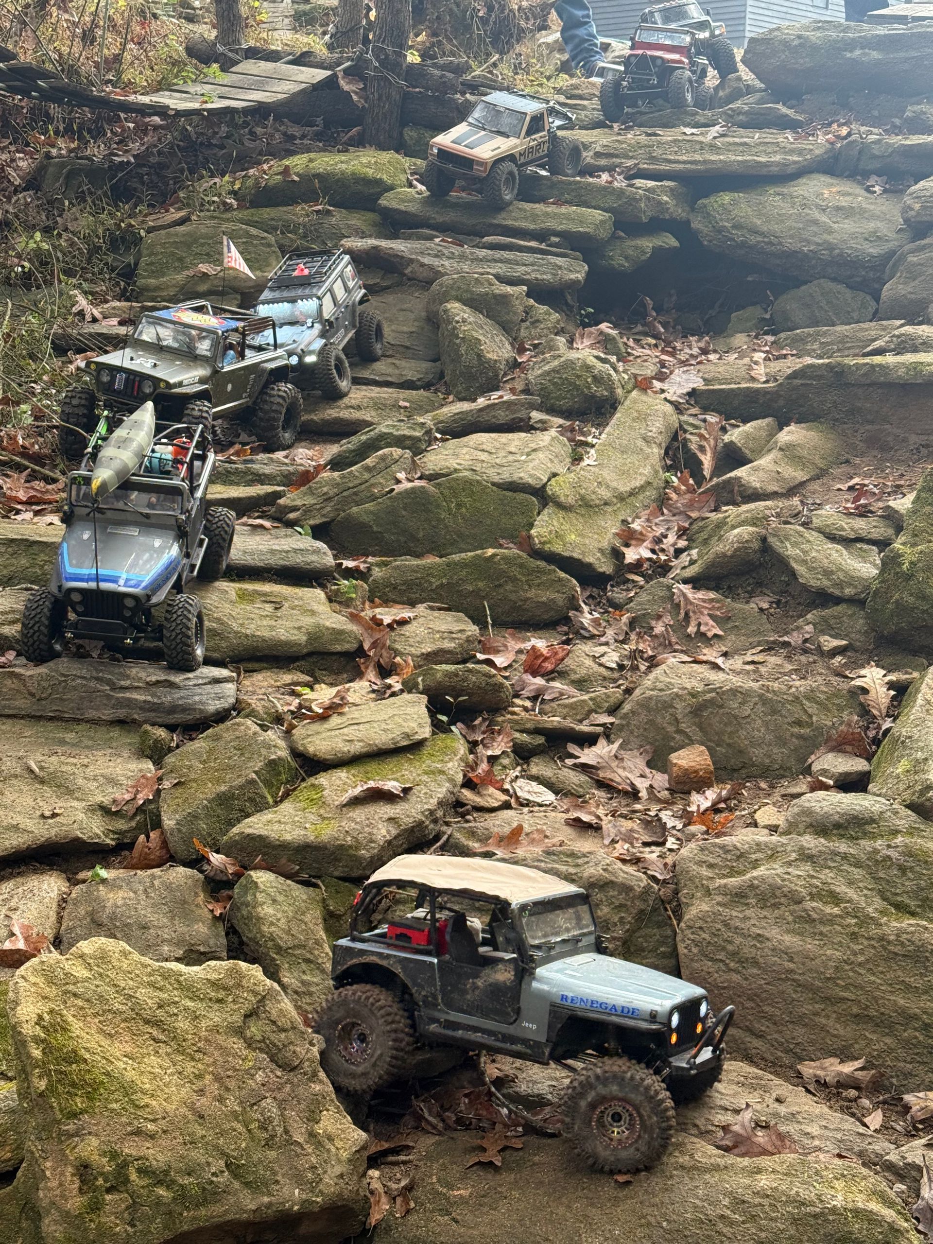 Remote control vehicles climbing a rocky terrain. Several blue and gray toy Jeeps traverse a rock-filled pathway outdoors.
