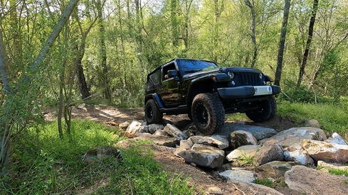 A black jeep is driving down a rocky trail in the woods.