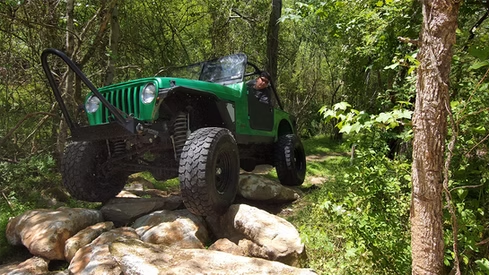 A green jeep is driving through a rocky area in the woods.
