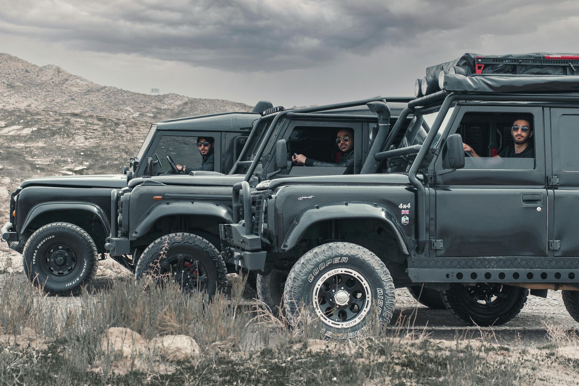 Three black Land Rover Defenders with occupants in a desert landscape.