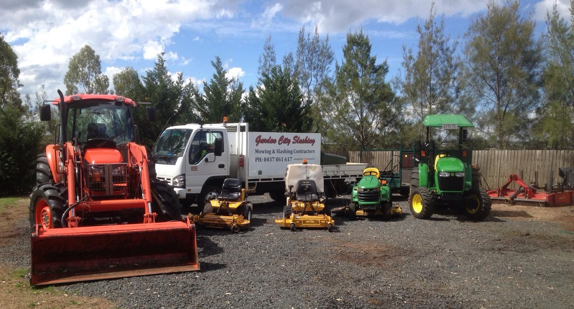 Orange tractor, white truck, and various lawn care equipment parked on gravel under a blue sky— Garden City Slashing in Highfields, QLD