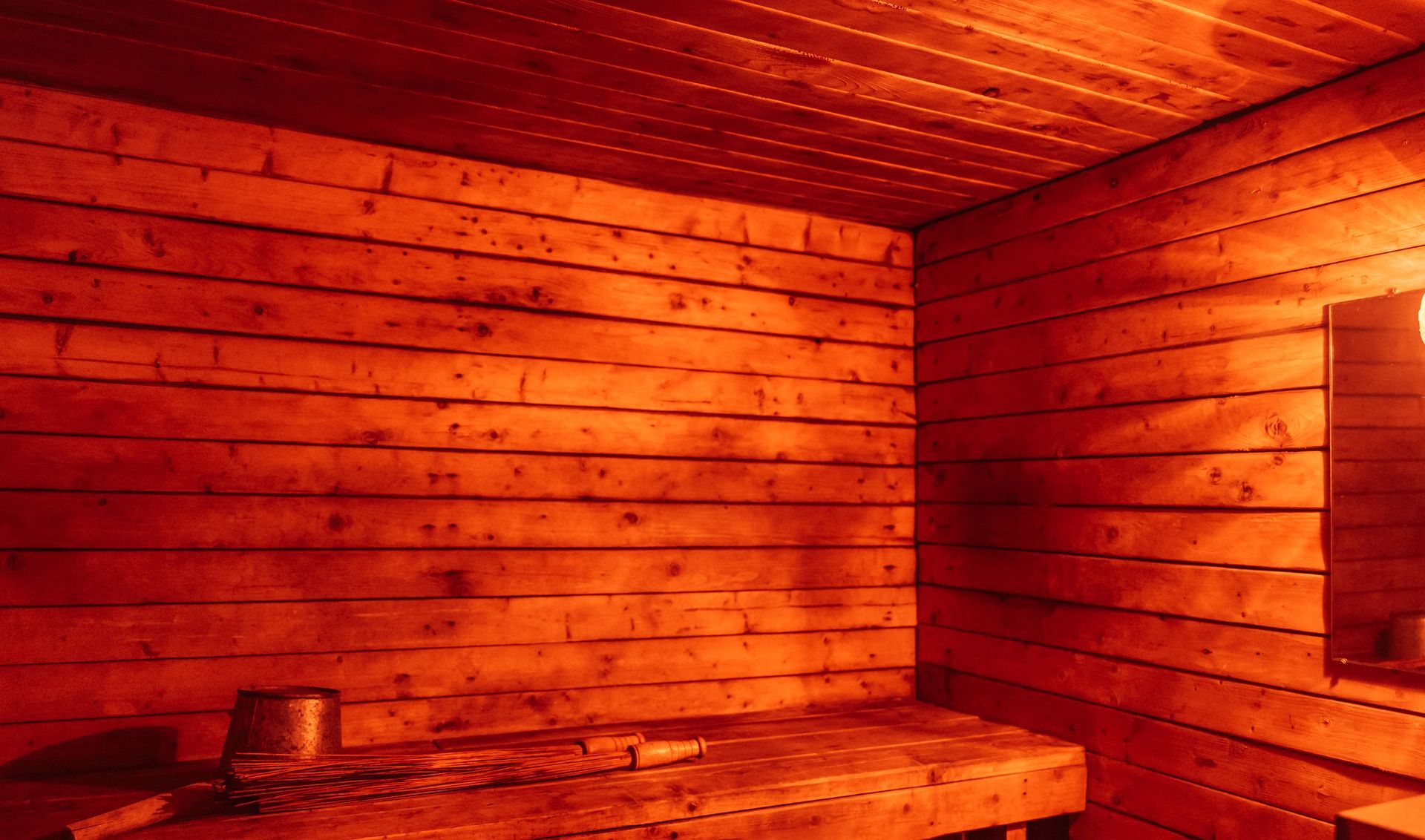 Wooden sauna interior, bathed in orange light. Bench, bucket, and mirror visible. Wooden sauna interior, bathed in orange light. Bench, bucket, and mirror visible.