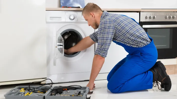 A man is kneeling on the floor fixing a washing machine.