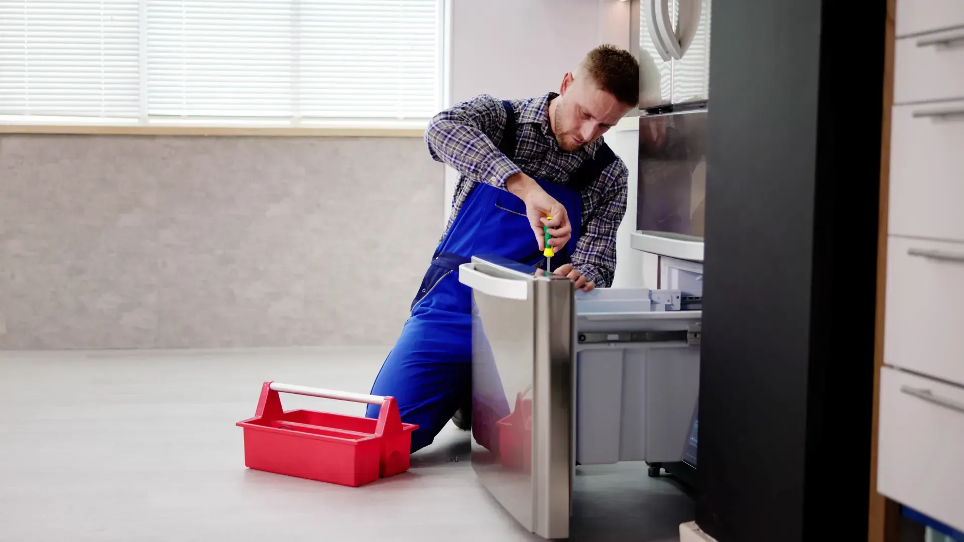 A man is kneeling on the floor fixing a refrigerator.