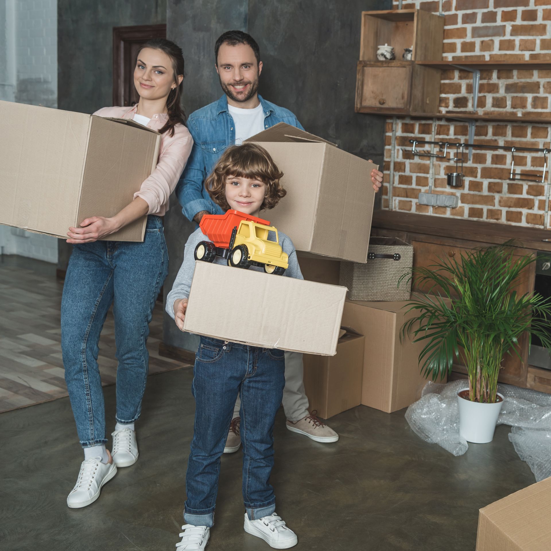 A family is moving into a new home and holding cardboard boxes and a toy truck.