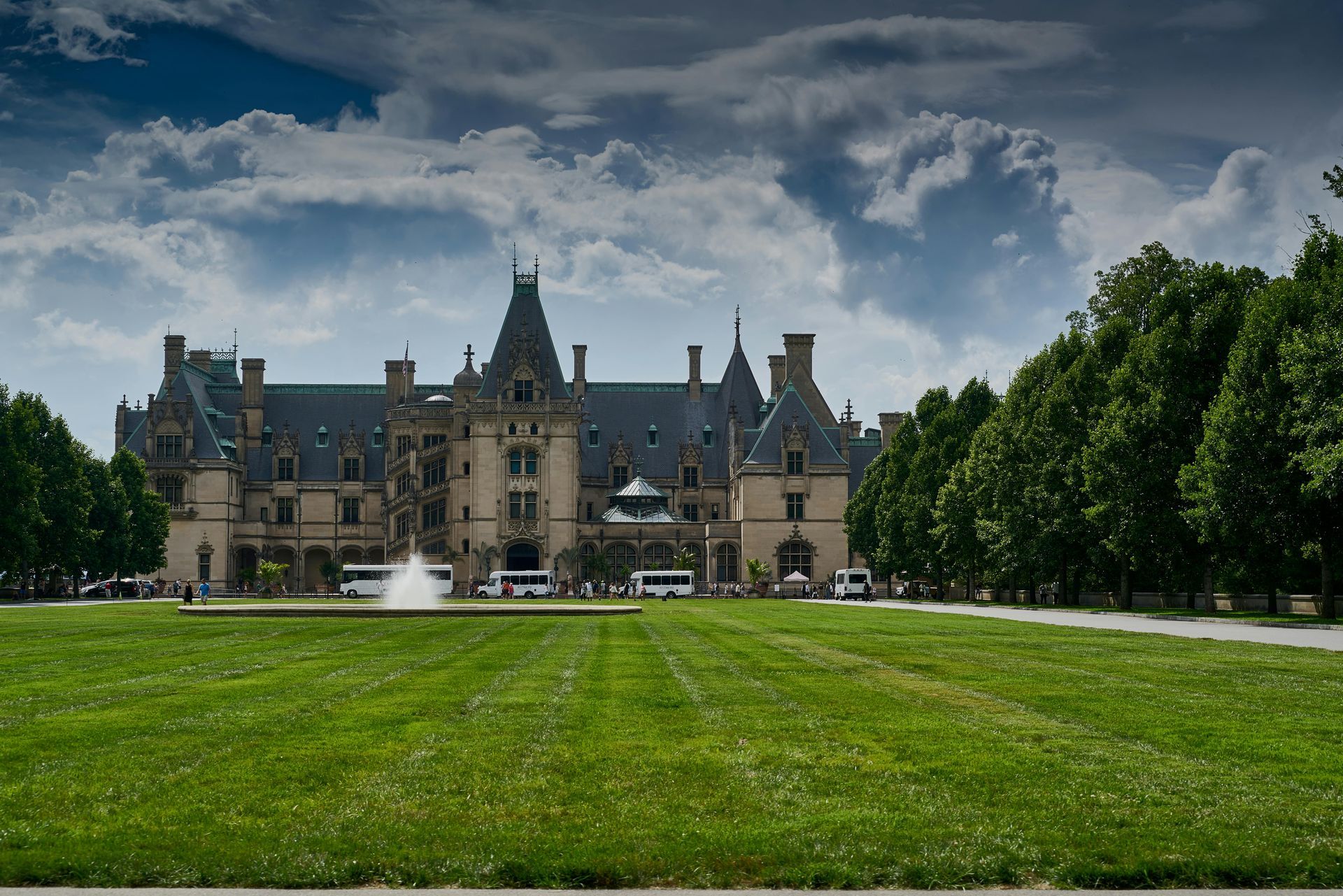 A large castle with a fountain in front of it is surrounded by trees and grass.