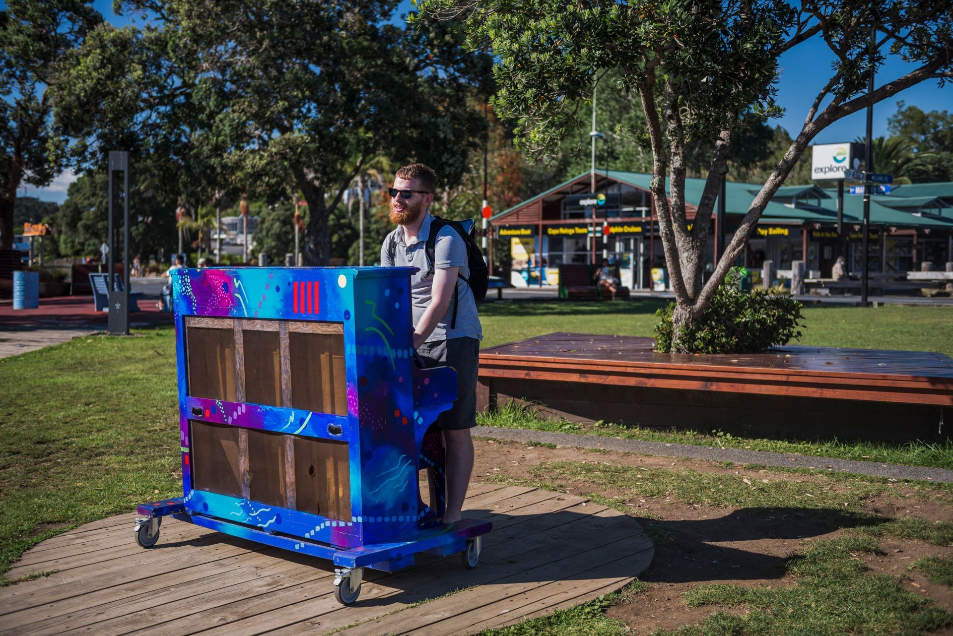 A man is pushing a blue cart with boxes on it in a park.