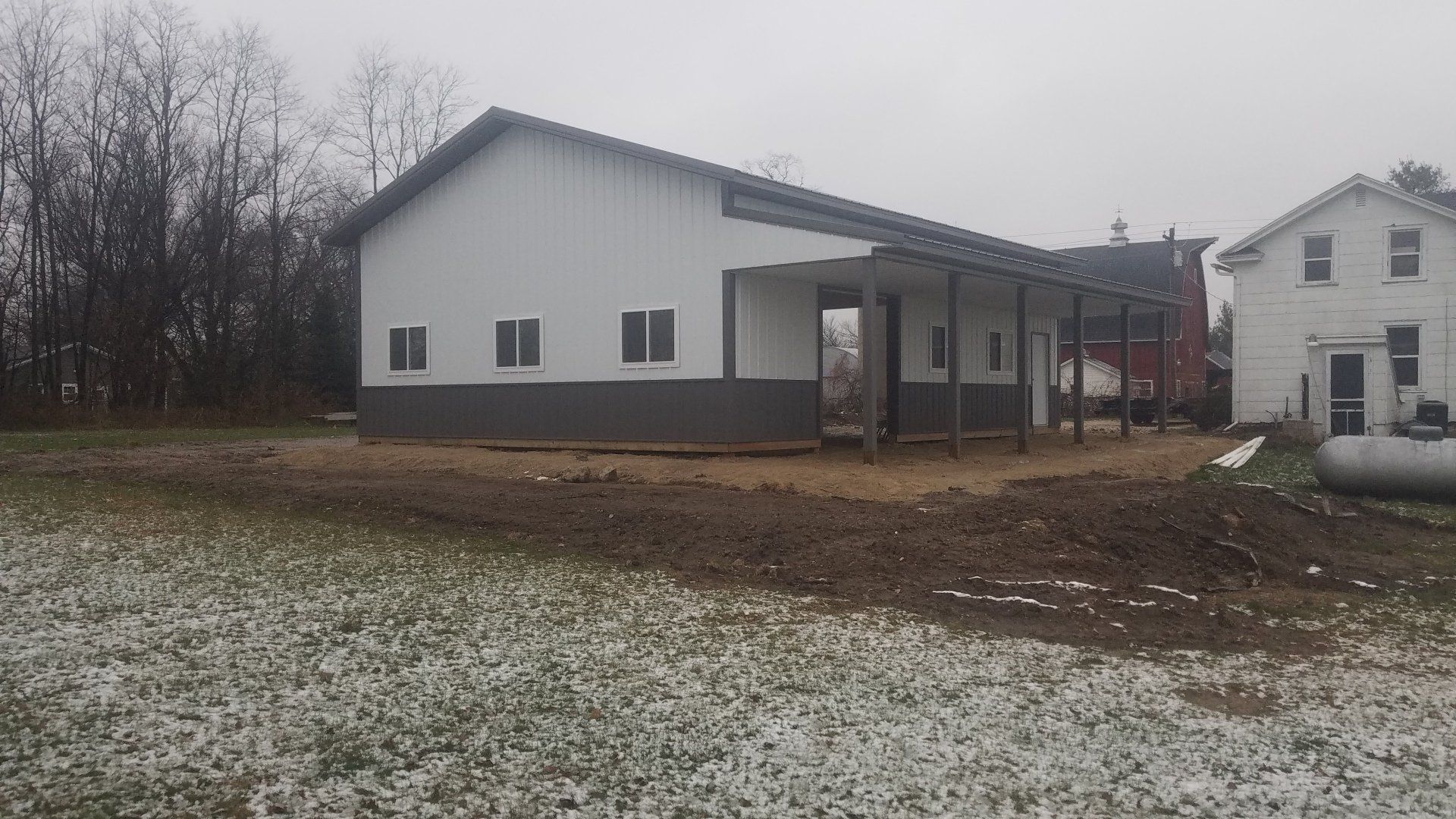 White barn with dark gray base and porch, in a field, with a white house in the background.