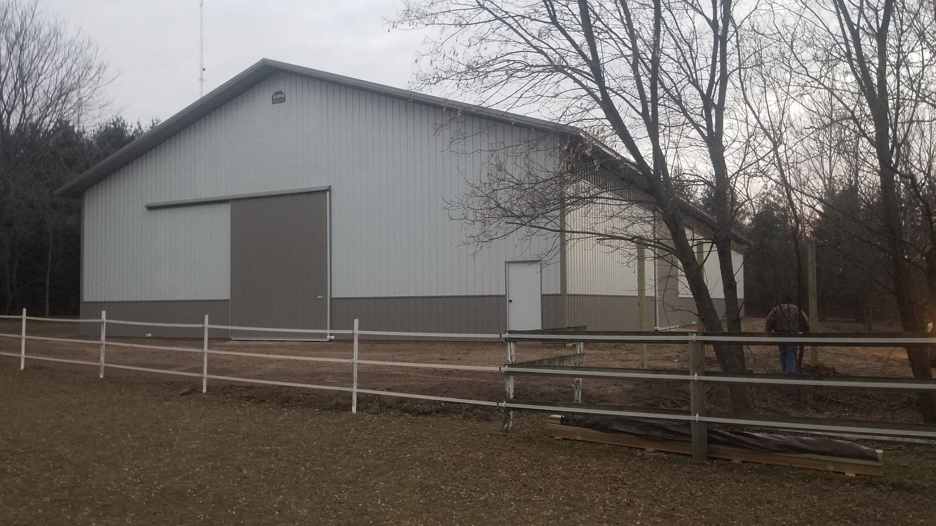 Large white and gray barn with a sliding door, next to a fence and trees.