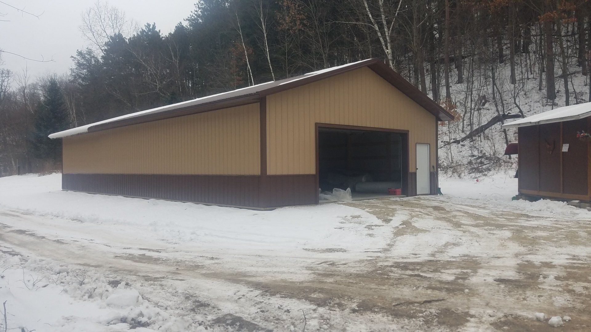Beige and brown metal shed in a snowy setting with an open garage door and a forest background.