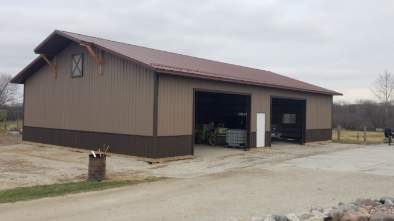 Brown and tan pole barn with two garage doors and a brown roof, set on a gravel driveway.