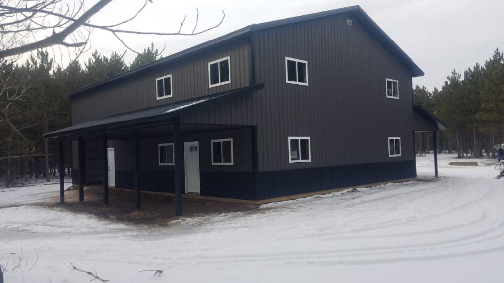 Dark gray metal building with white-trimmed windows and a covered porch in a snowy, wooded area.