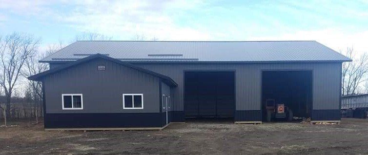 Dark gray metal building with two garage doors and a smaller attached section with two windows.