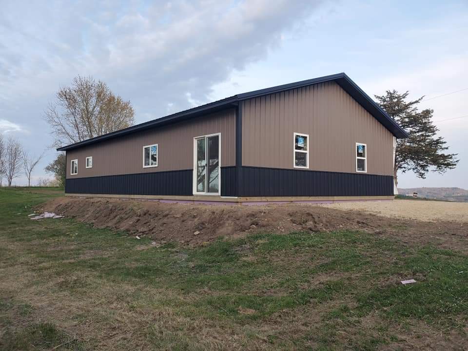 Tan and black metal building with white-framed windows and a sliding glass door, on a grassy hillside.