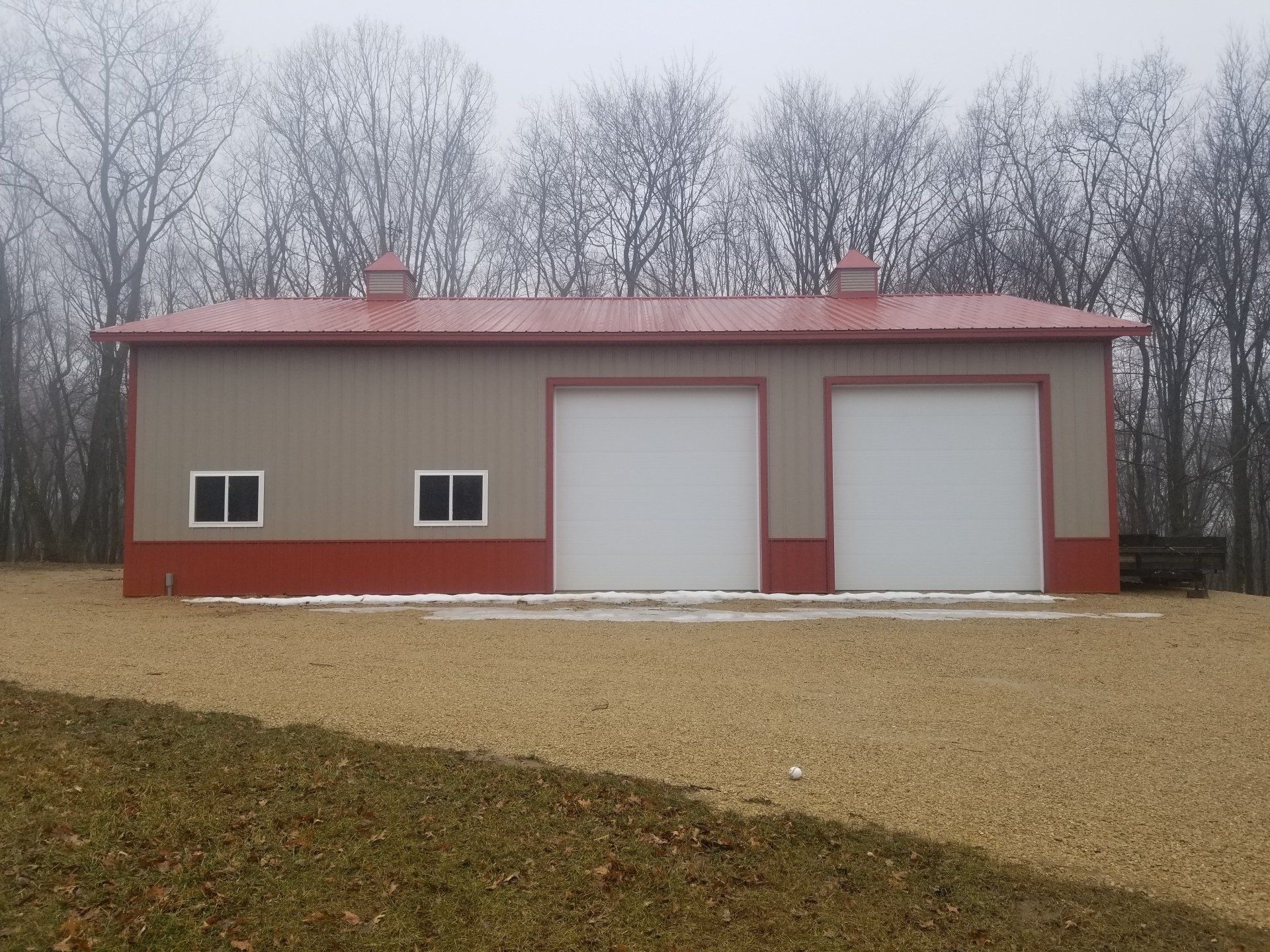 Tan and red metal garage with two bays, set in a gravel yard, with trees in the background.