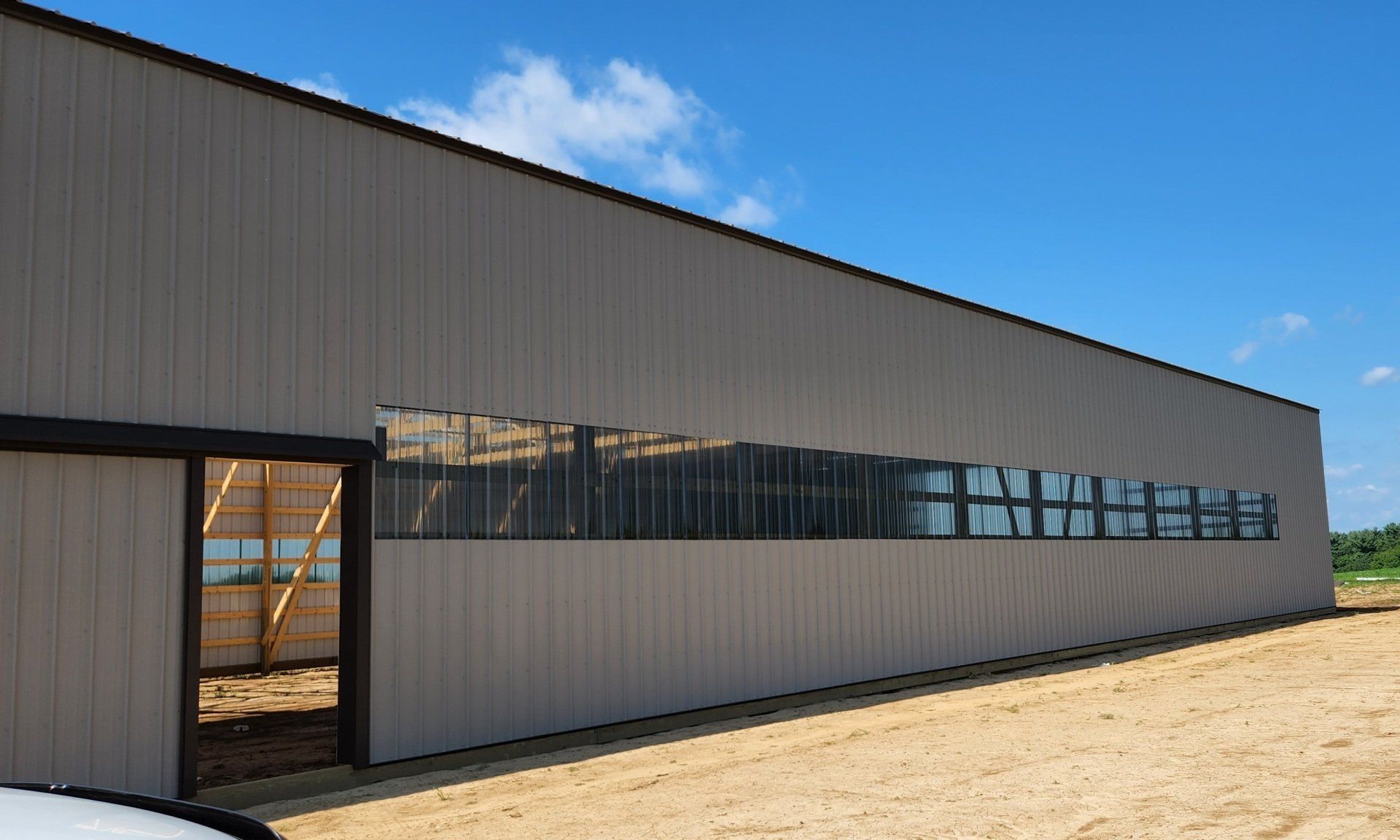 Beige metal building with a long row of windows, a door open to a wooden frame, and a clear blue sky.