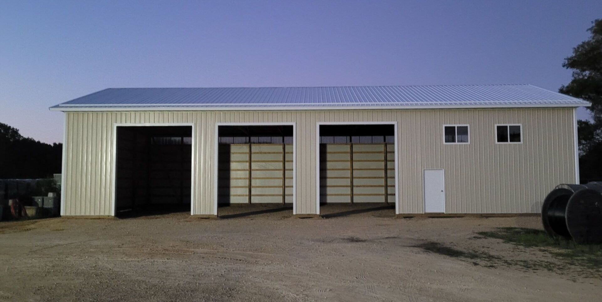 A large metal building with three garage bays. Gravel yard, trees, and dusk sky.