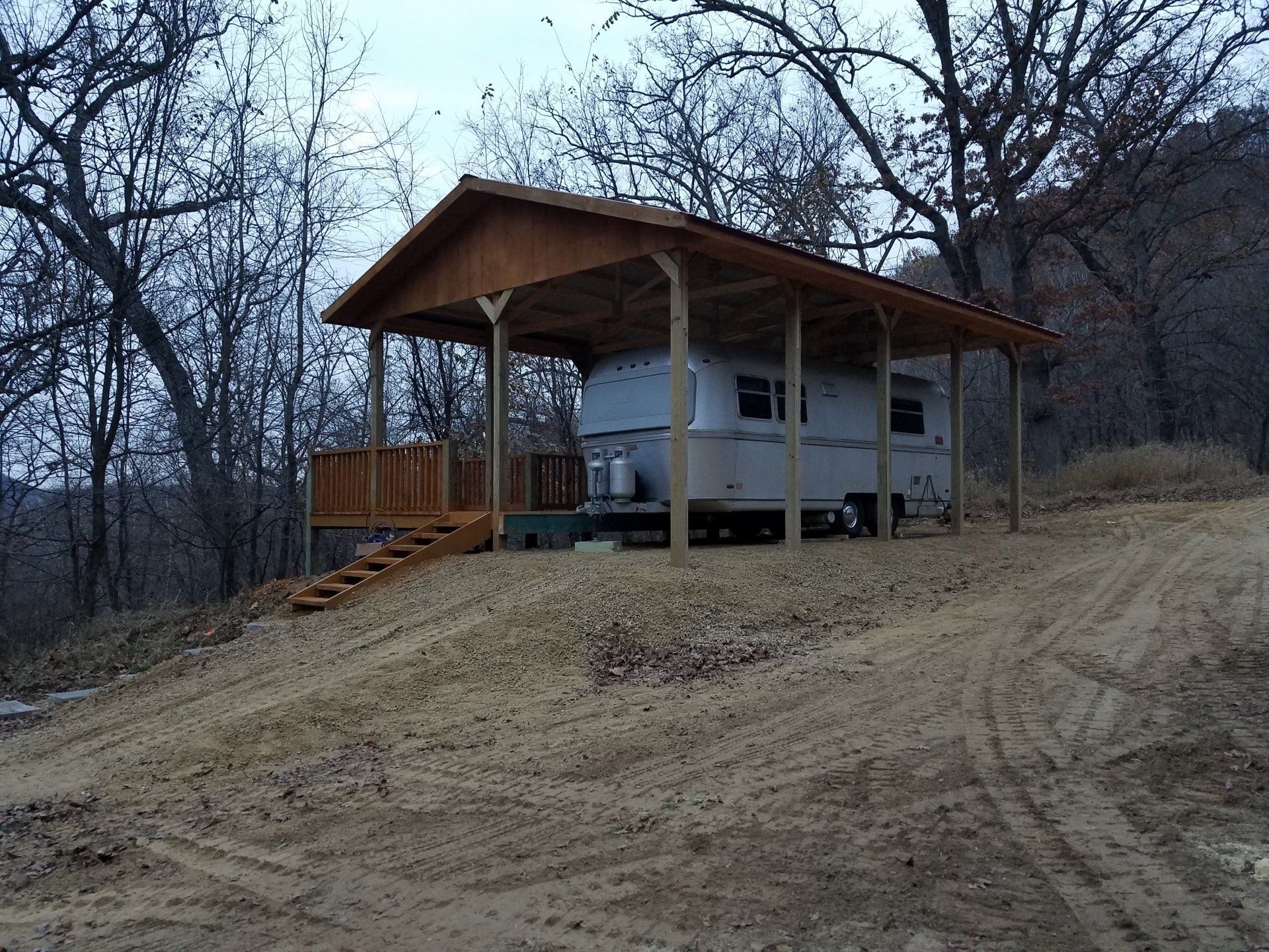 A vintage silver camper under a wooden roof on a dirt hill, steps lead to a porch.