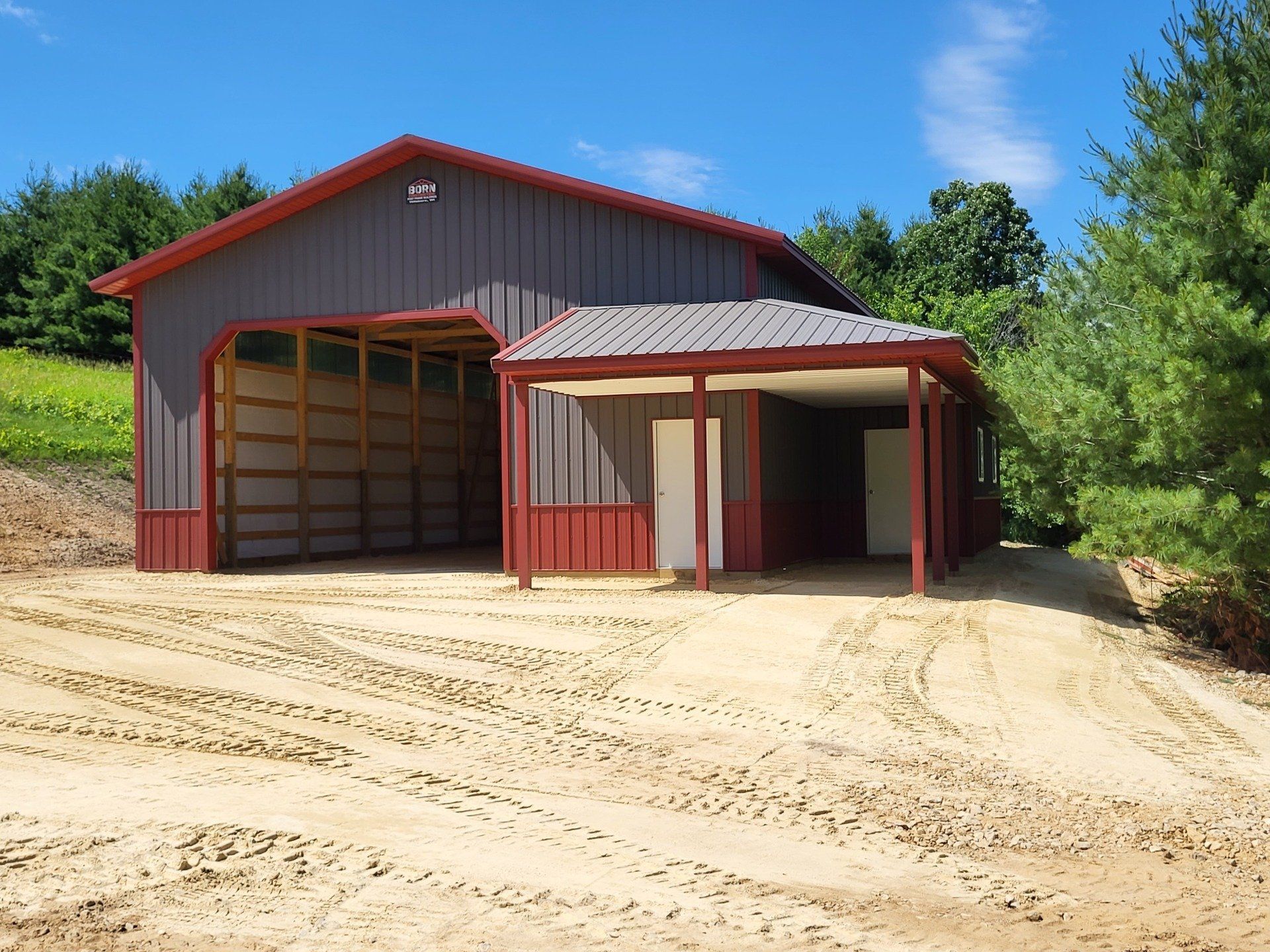 Gray and red barn with open bay and covered entry on a sandy drive, with green trees.