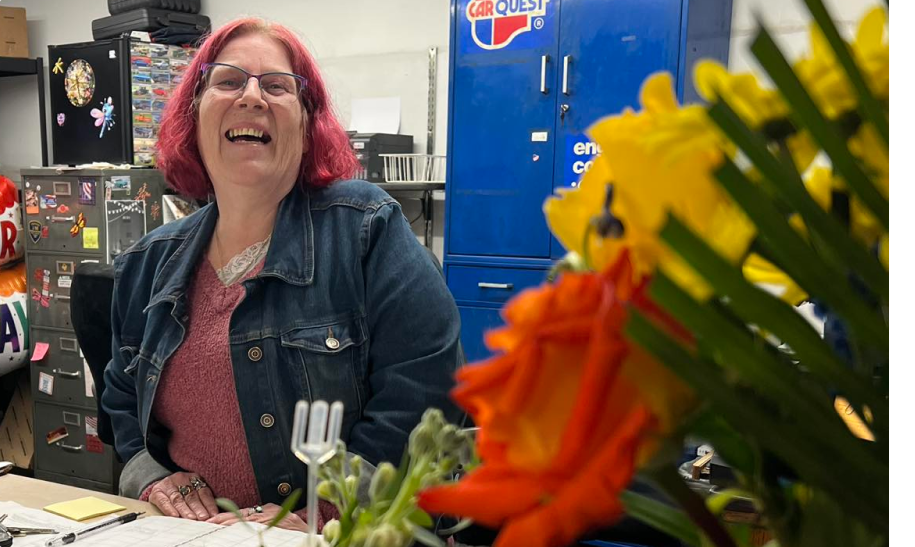 A woman with red hair is sitting at a desk in front of a vase of flowers.