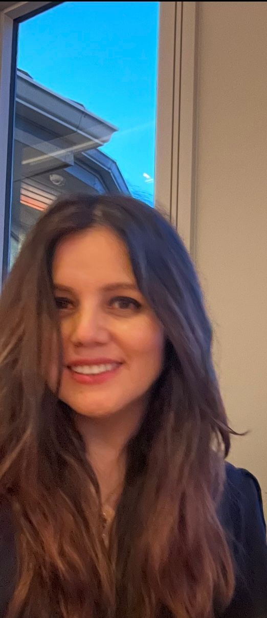 Smiling woman with long brown hair in front of a window, wearing a dark top