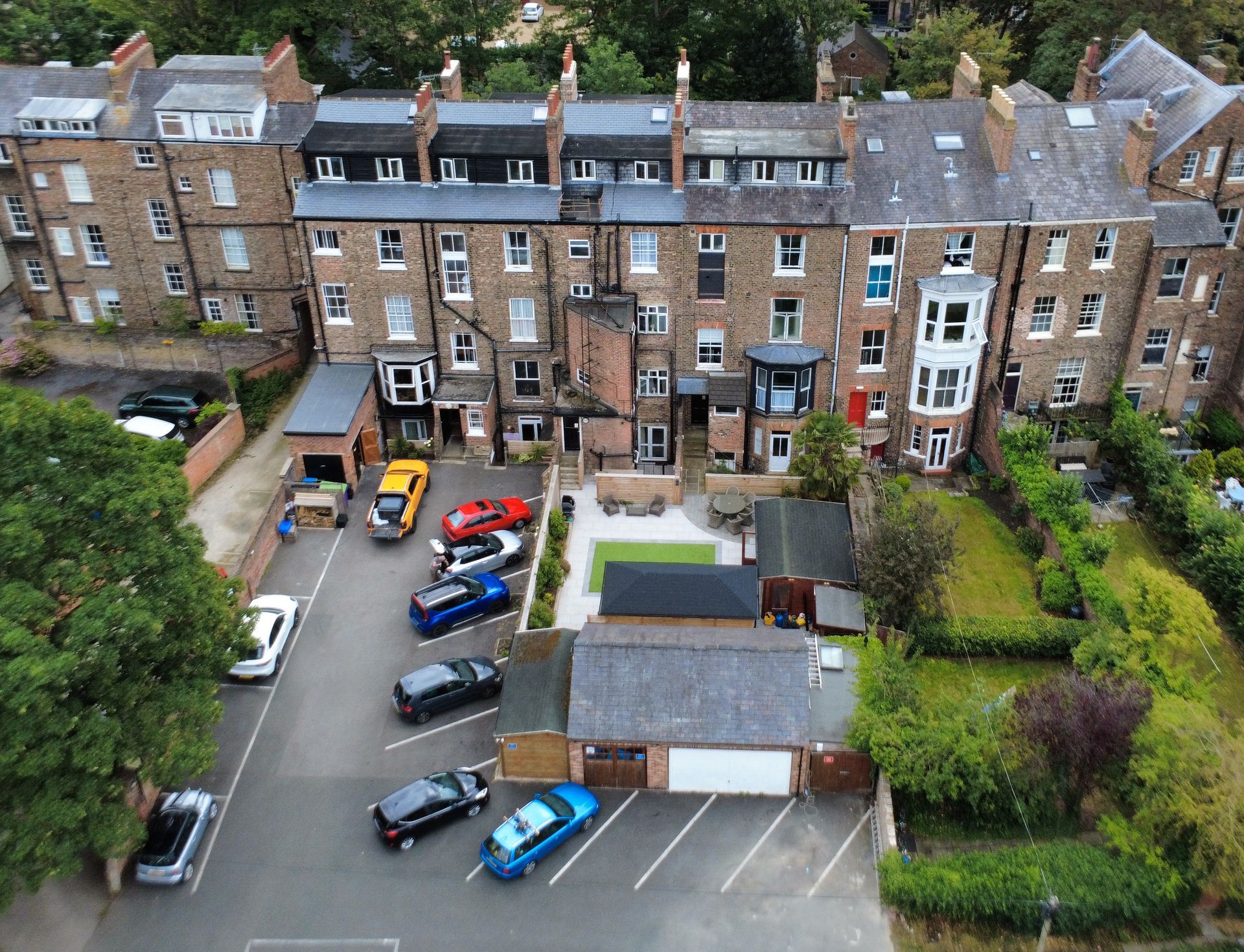 Aerial view of a row of brick buildings with cars parked in front. Green trees and a small garden are also visible.