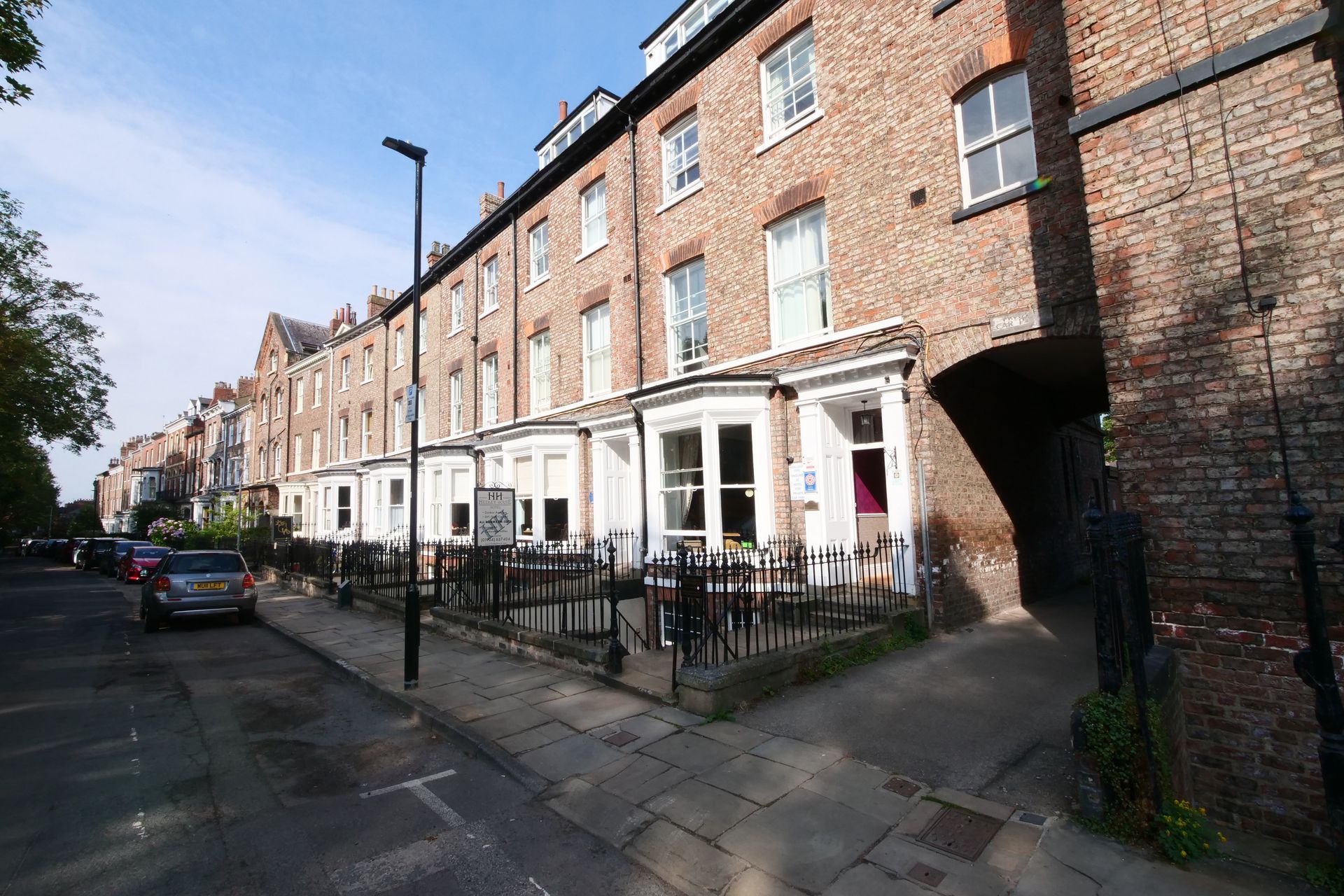 Row of brick buildings, one with a white-framed bay window and tables outside. Cars line the street.